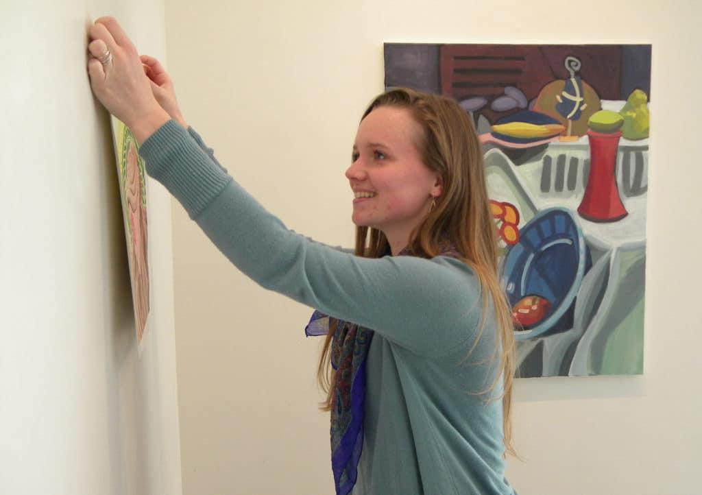 Woman hanging a picture on a white wall with colorful artwork from Howard Payne University in the background. | HPU